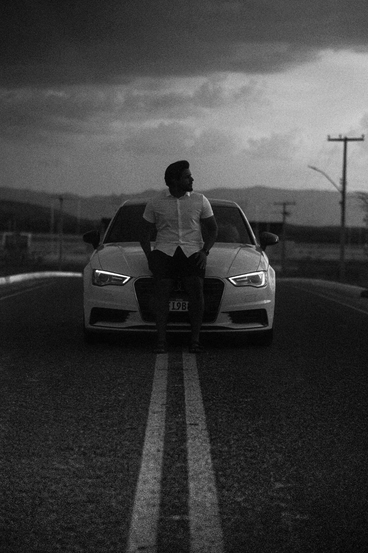Micael Brito Monteiro encostado num carro branco no meio de uma estrada, em preto e branco